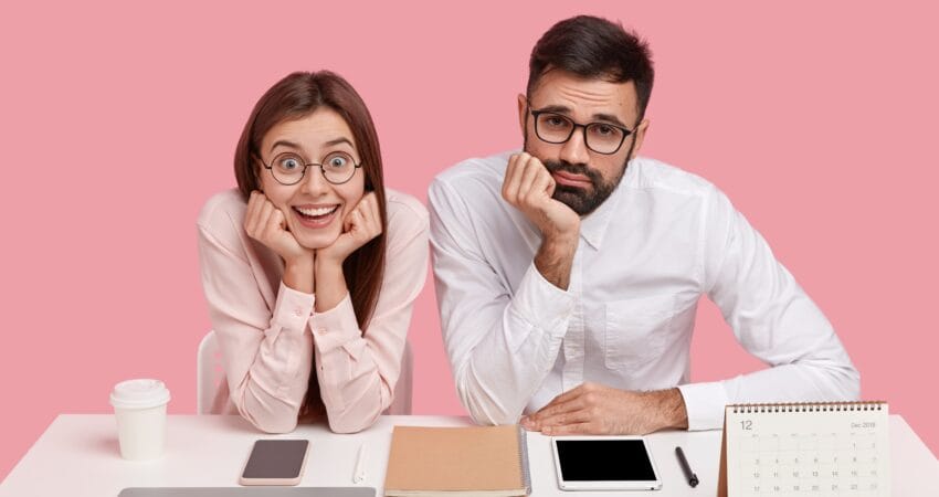 Two office employees sitting side by side at a desk with neutral and disengaged expressions, surrounded by work tools like a laptop, tablet, notebook, and calendar, illustrating workplace monotony and employee disengagement that can lead to attrition.