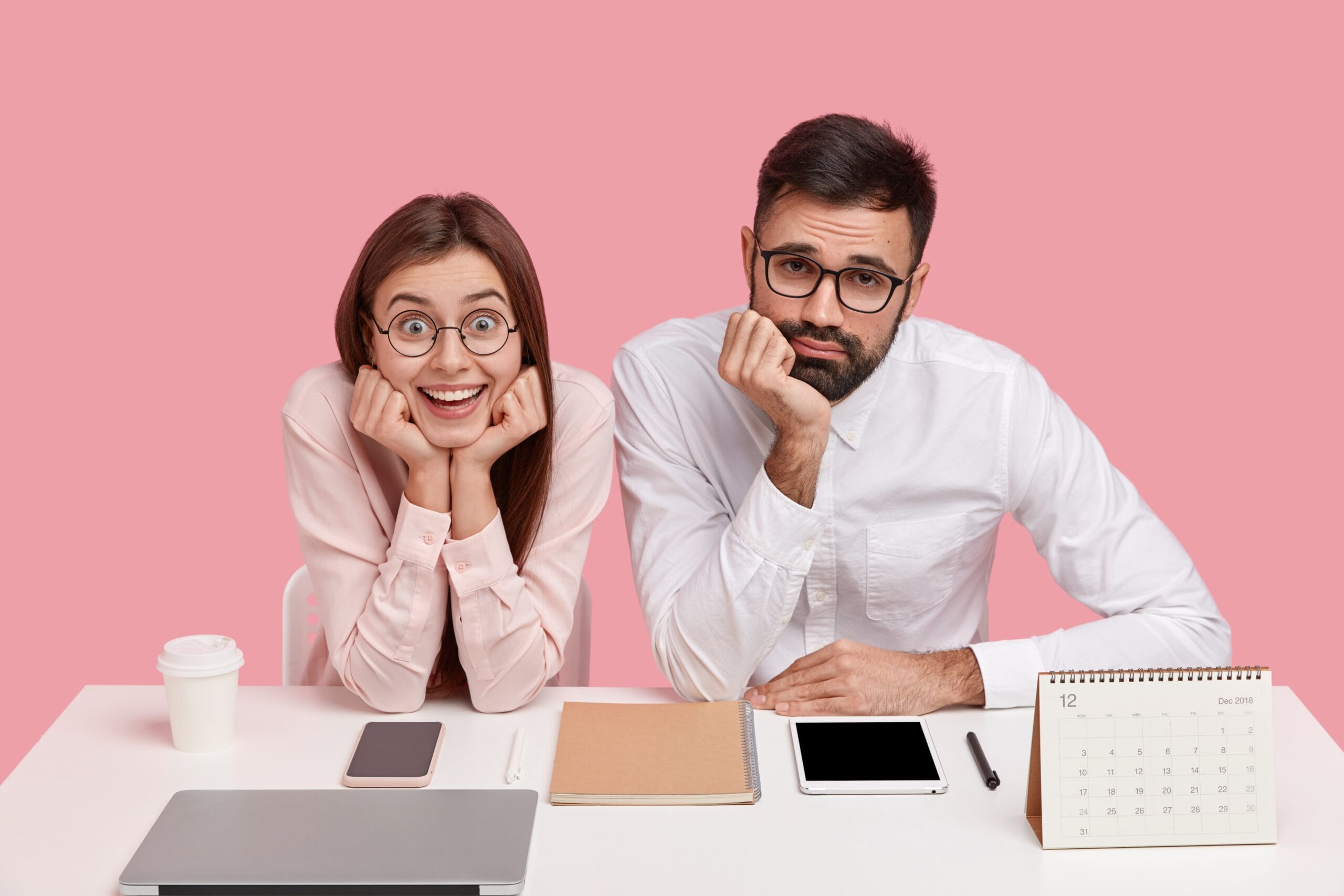 Two office employees sitting side by side at a desk with neutral and disengaged expressions, surrounded by work tools like a laptop, tablet, notebook, and calendar, illustrating workplace monotony and employee disengagement that can lead to attrition.
